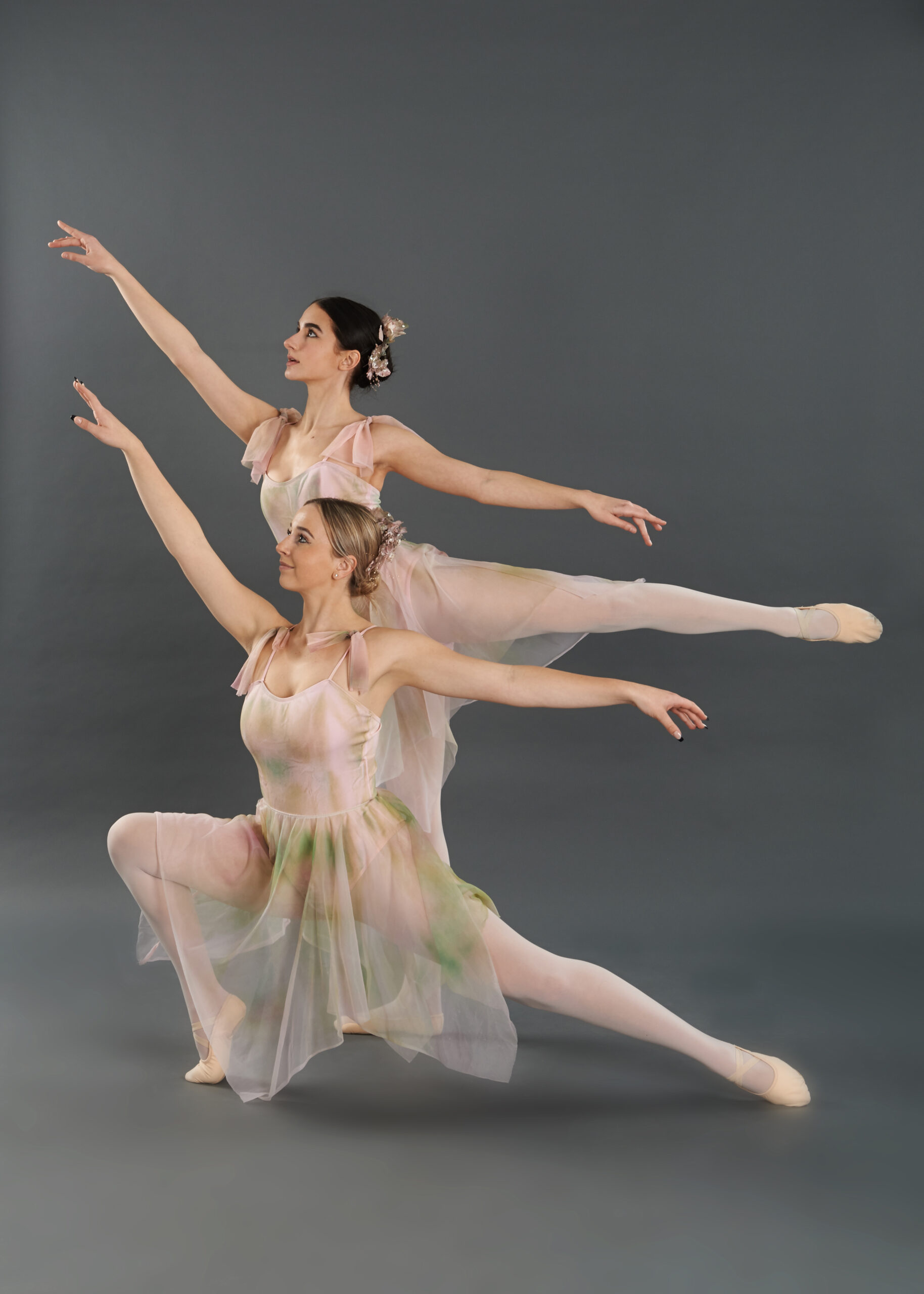 Two female dancers in costume in similar pose with one in lower position in front of other and arms outstretched