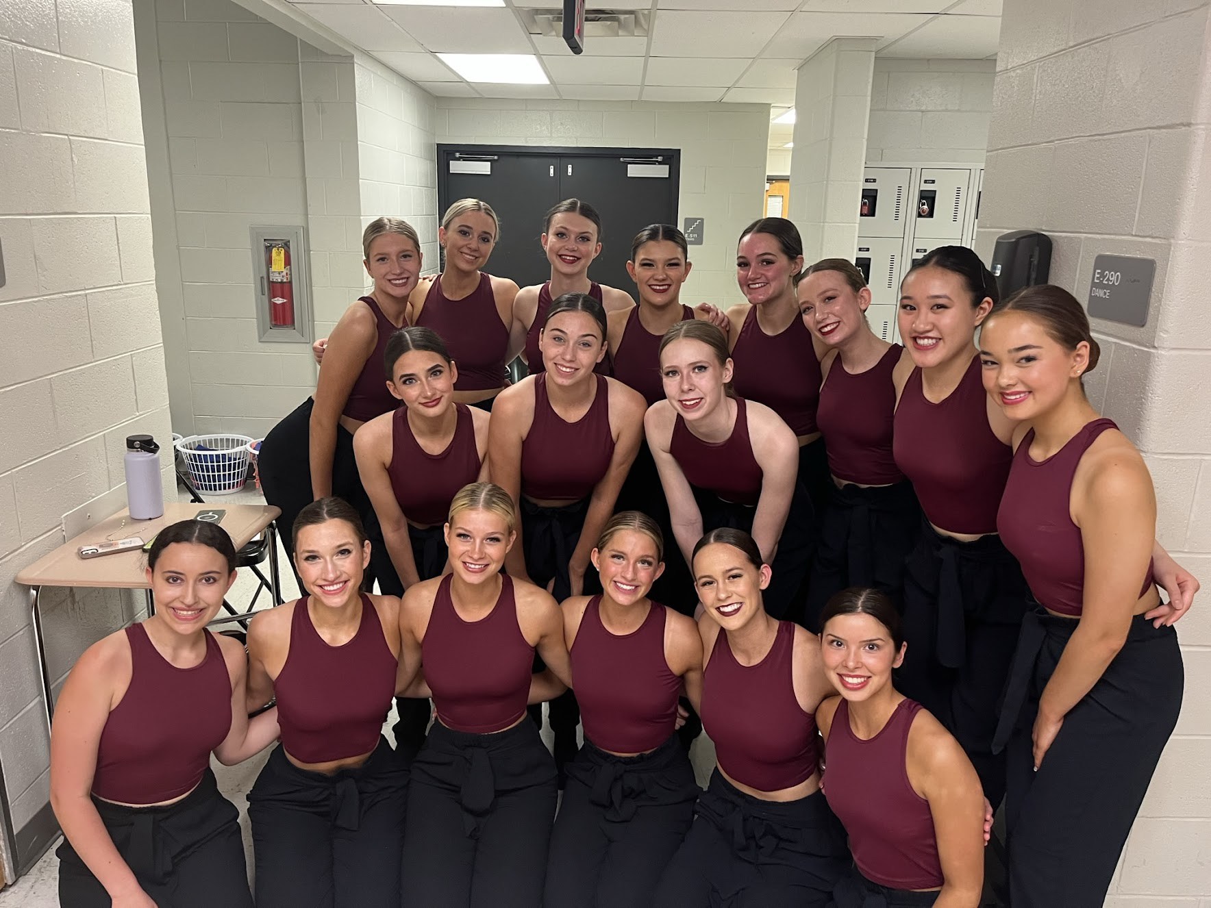 Female dancers wearing maroon and black matching dance costumes in three rows with arms wrapped around each other while smiling and facing forward